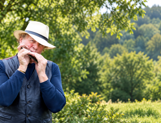 Musician wearing a white fedora and blue shirt plays the harmonica outdoors against a backdrop of green trees.