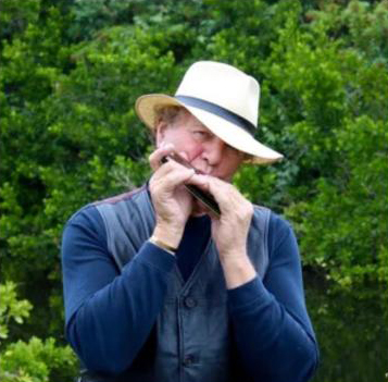 Musician wearing a white fedora and blue shirt plays the harmonica outdoors against a backdrop of green trees.